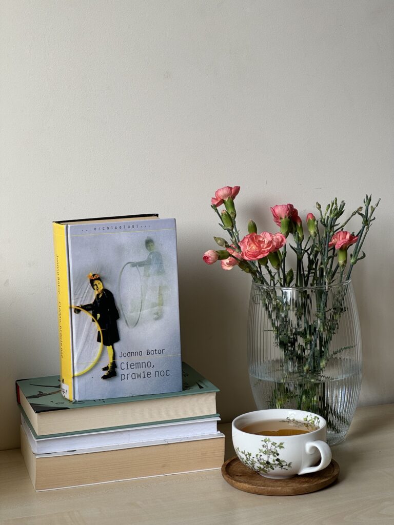 Kolorowa fotografia. Na stole stoik książek. Na ich szczycie stoi książka Ciemno, prawie noc. Obok wazon z goździkami oraz filiżanka z herbatą.