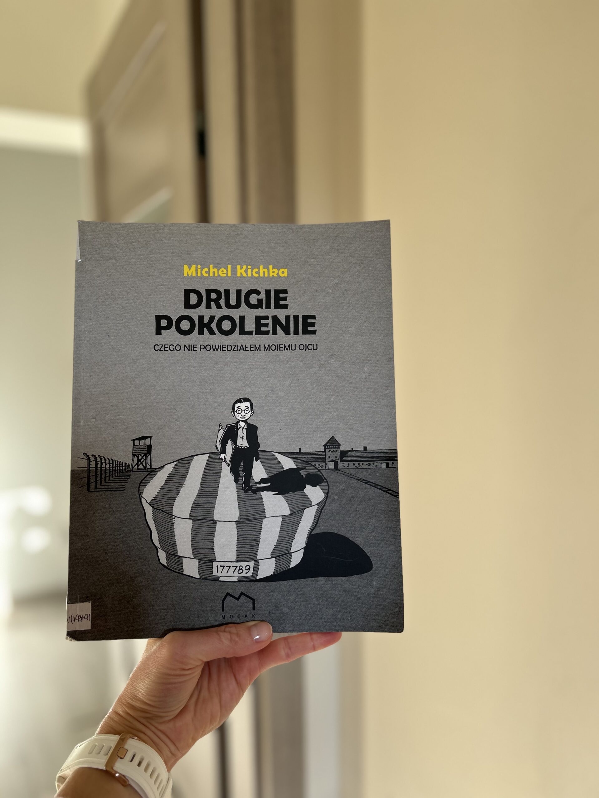 Kolorowa fotografia. Wyciągnięta dłoń trzyma książkę. W tle otwarte drzwi.