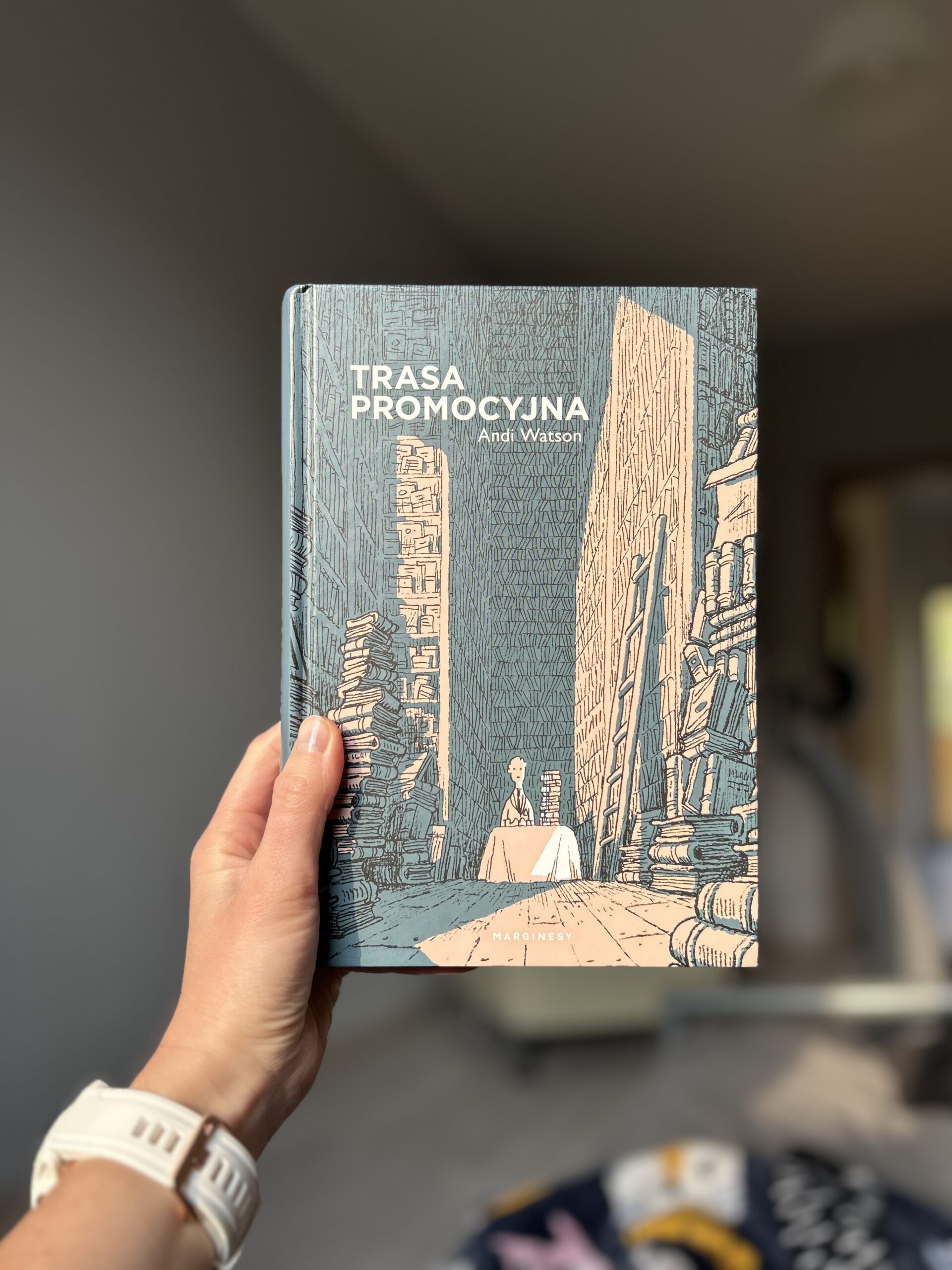 Kolorowa fotografia. Wyciągnięta dłoń trzyma książkę. W tle pokój w mieszkaniu.