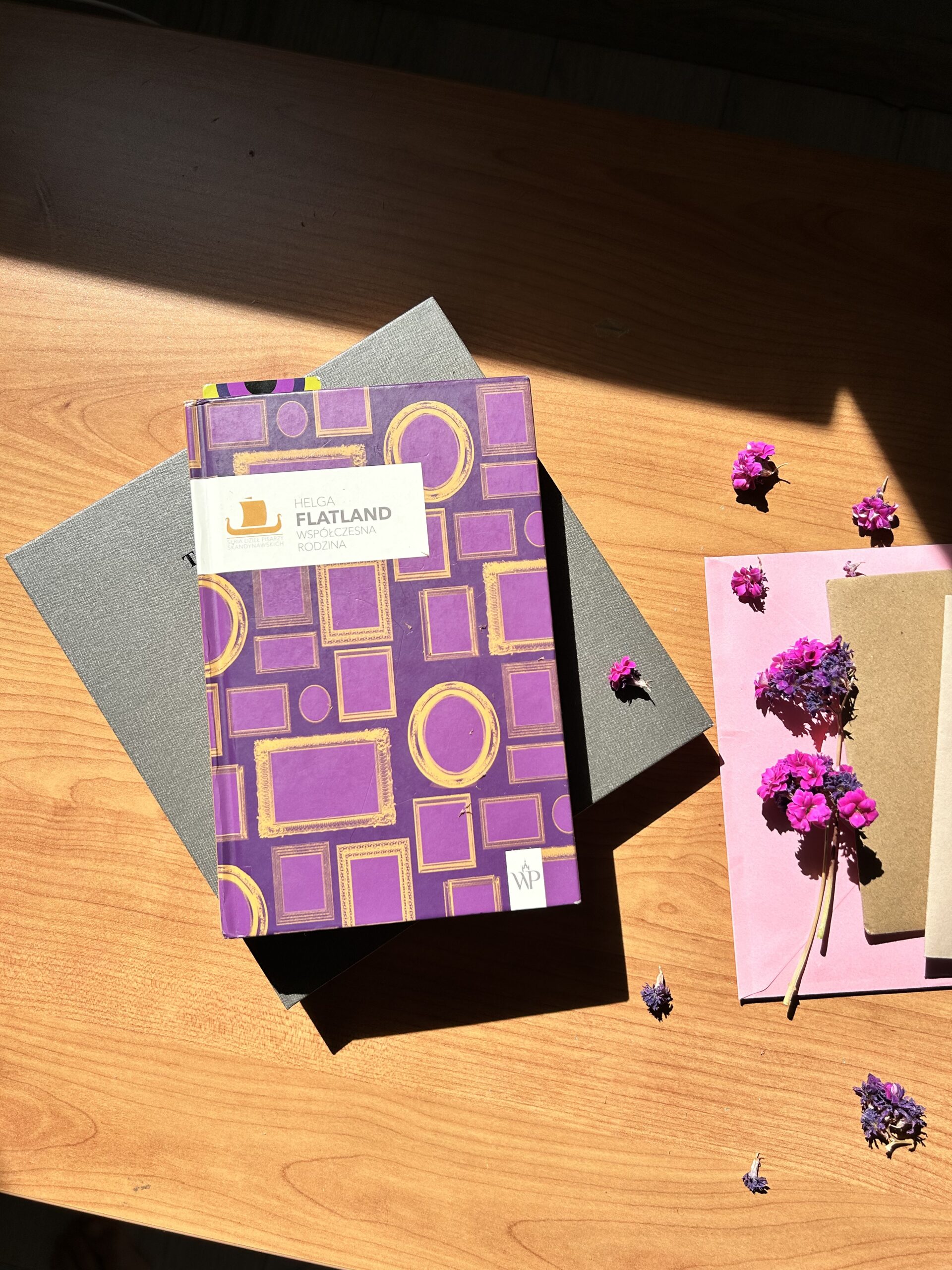 Kolorowe zdjęcie. Na stole leży książka. Obok kolorowe koperty. Wokół suszone kwiaty o fioletowych płatkach.