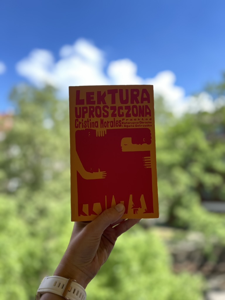 Kolorowa fotografia. Wyciągnięta kobieca dłoń trzyma książkę. W tle błękitne niebo i drzewa.