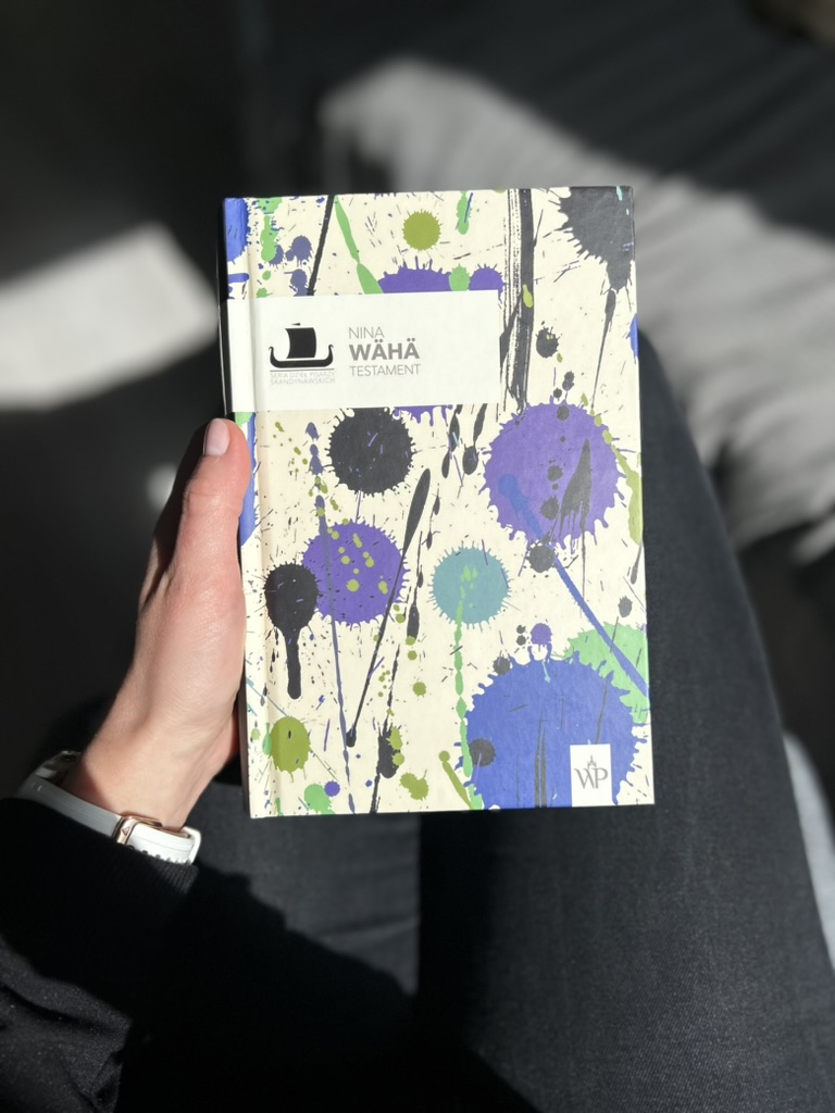 Kolorowa fotografia. Kobieca ręka trzyma książkę na kolanach. W tle szara narzuca na łóżku.