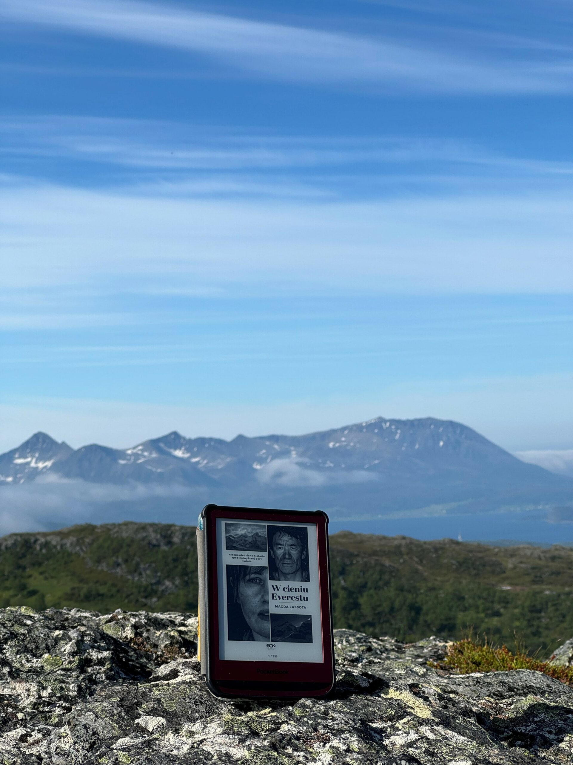 Kolorowe zdjęcie. Czytnik e-booków leży na kamieniu. W tle pasmo górskie. Na ekranie czytnika okładka książki.