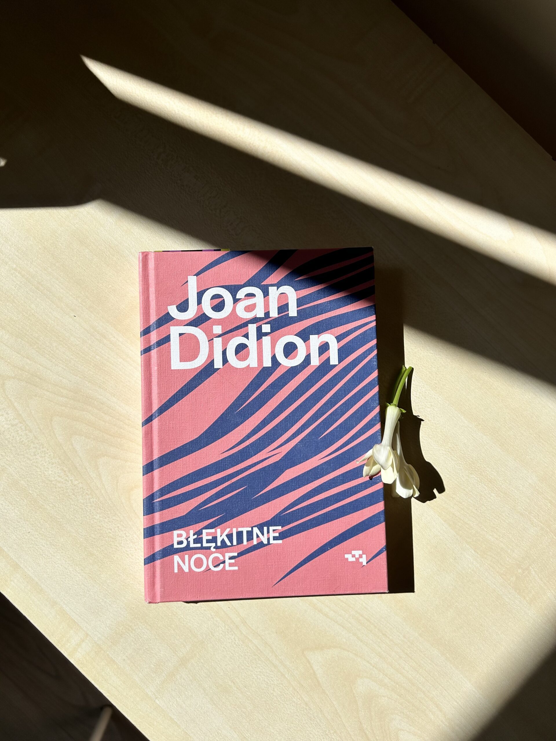 Kolorowe zdjęcie. Książka leży na biurku. Obok leżą 2 białe kwiaty stefanotisa.