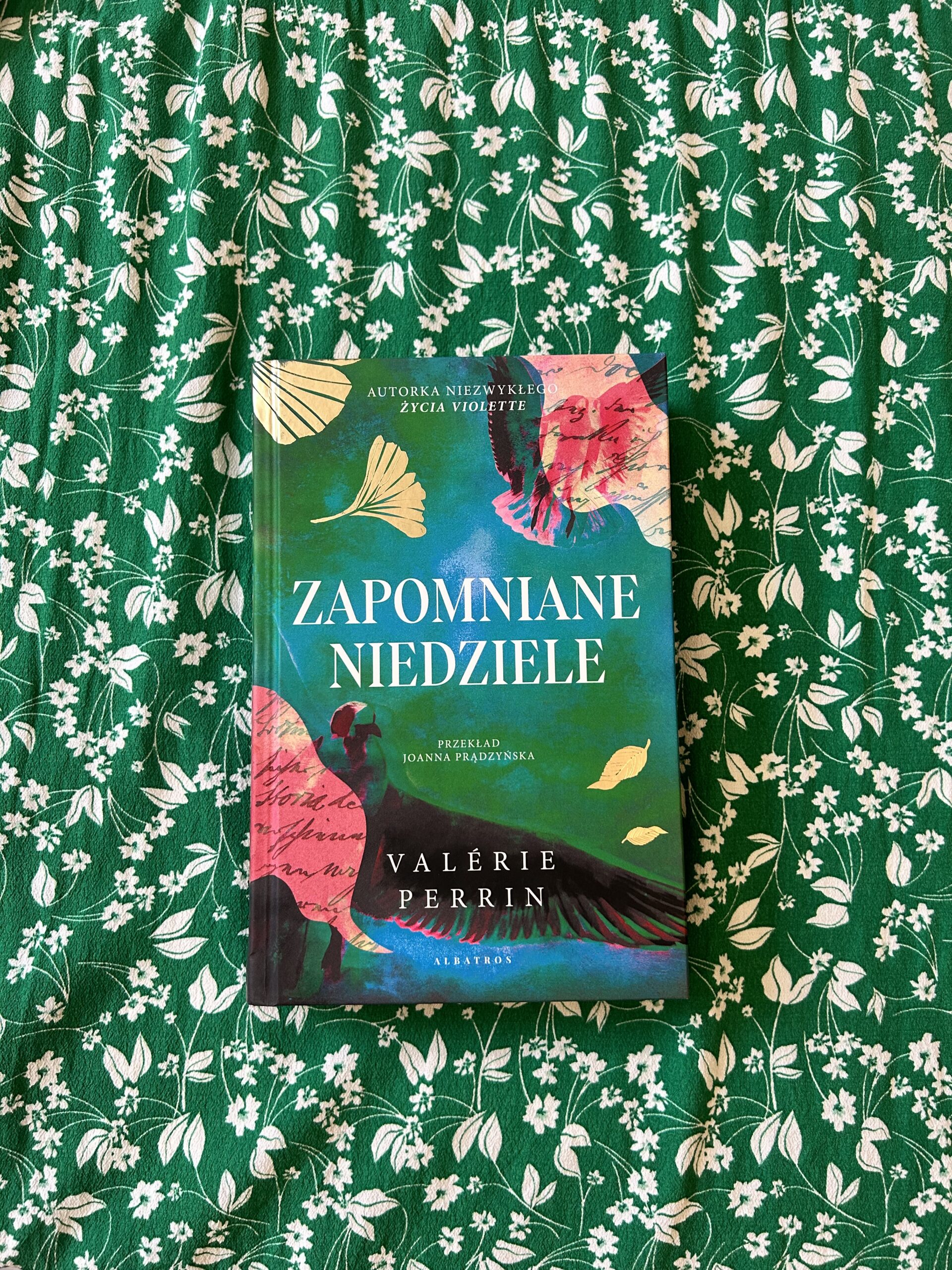 Kolorowe zdjęcie. Książka leży na stole przykrytym zielonym materiałem w drobne, białe kwiatki.