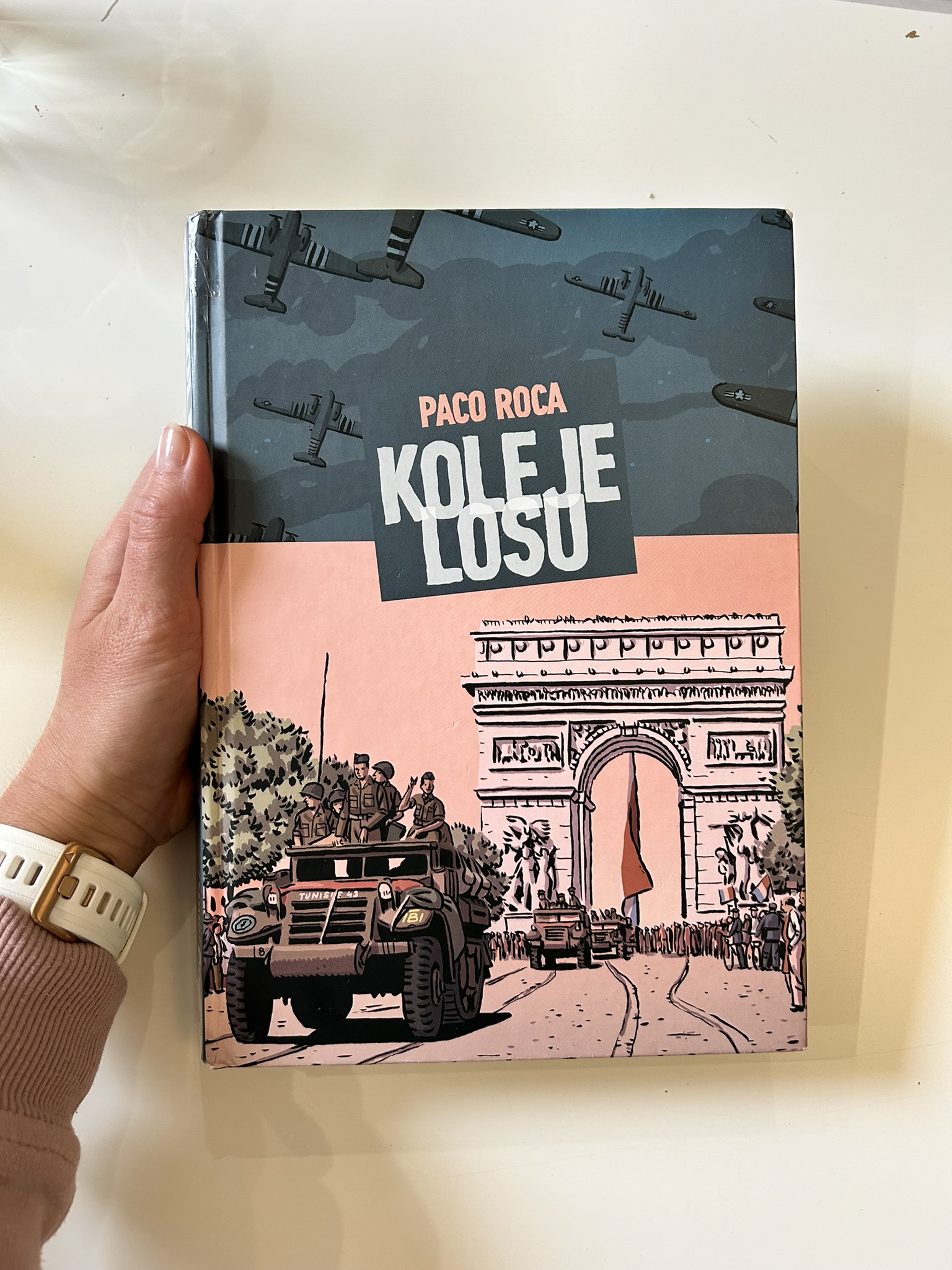 Kolorowa fotografia. Damska dłoń trzyma książkę w twardej okładce. W tle blat białego stołu,