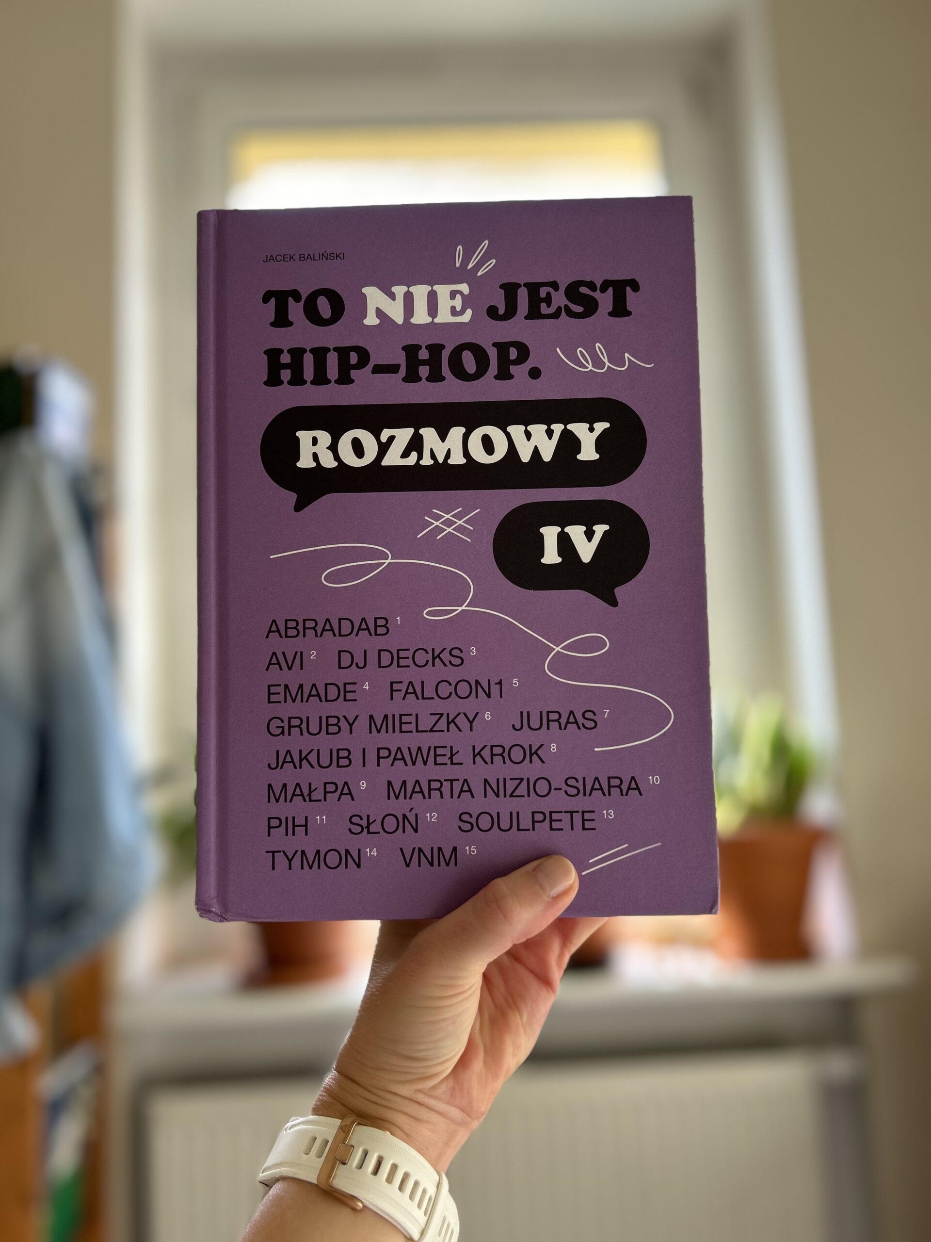 Kolorowe zdjęcie. Wyciągnięta ręka trzyma książkę. W tle okno i parapet z zielonymi roślinami w doniczkach.