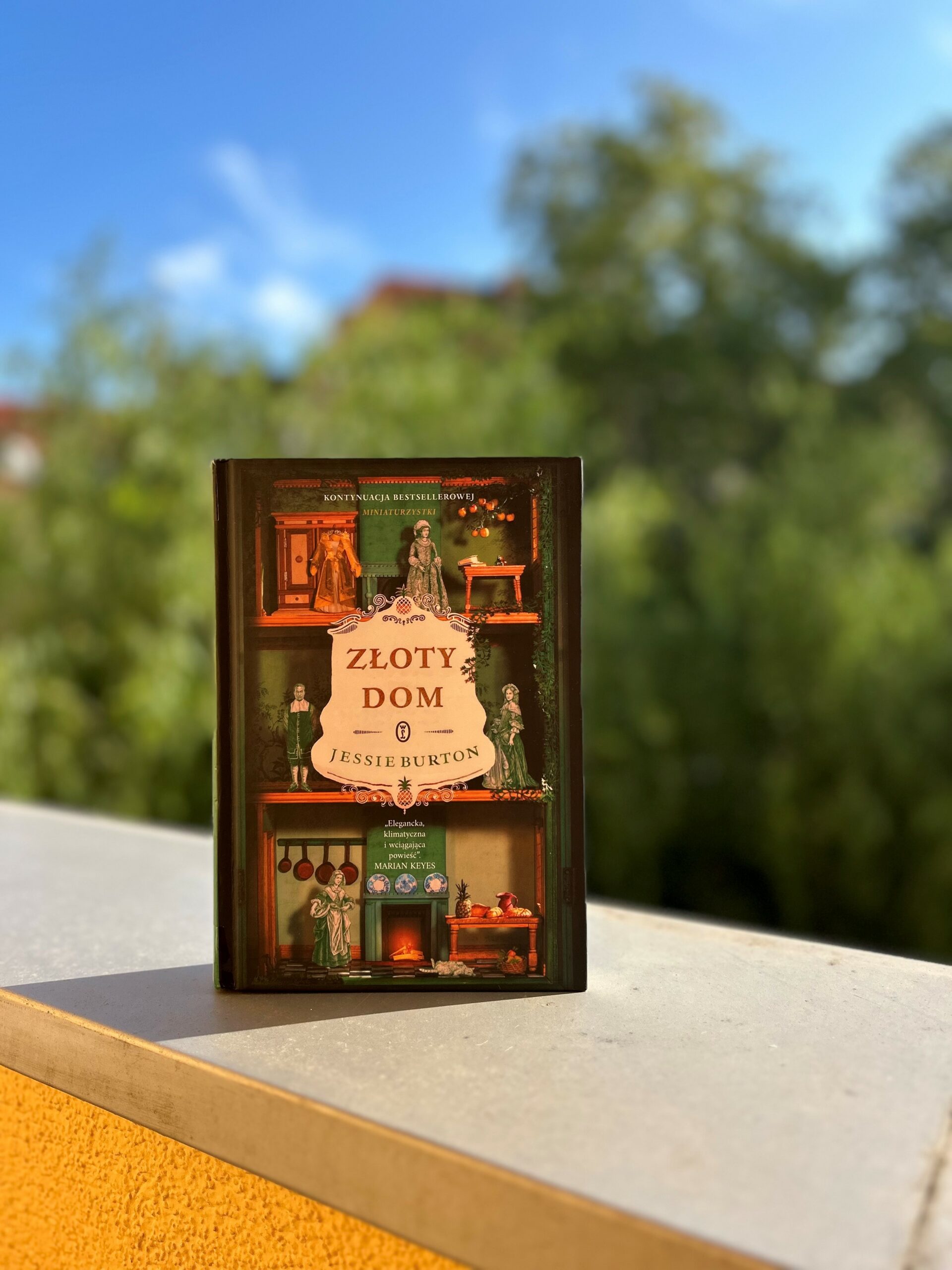 Kolorowe zdjęcie. Książka stoi na parapecie balkonu. W tle zielone drzewa.