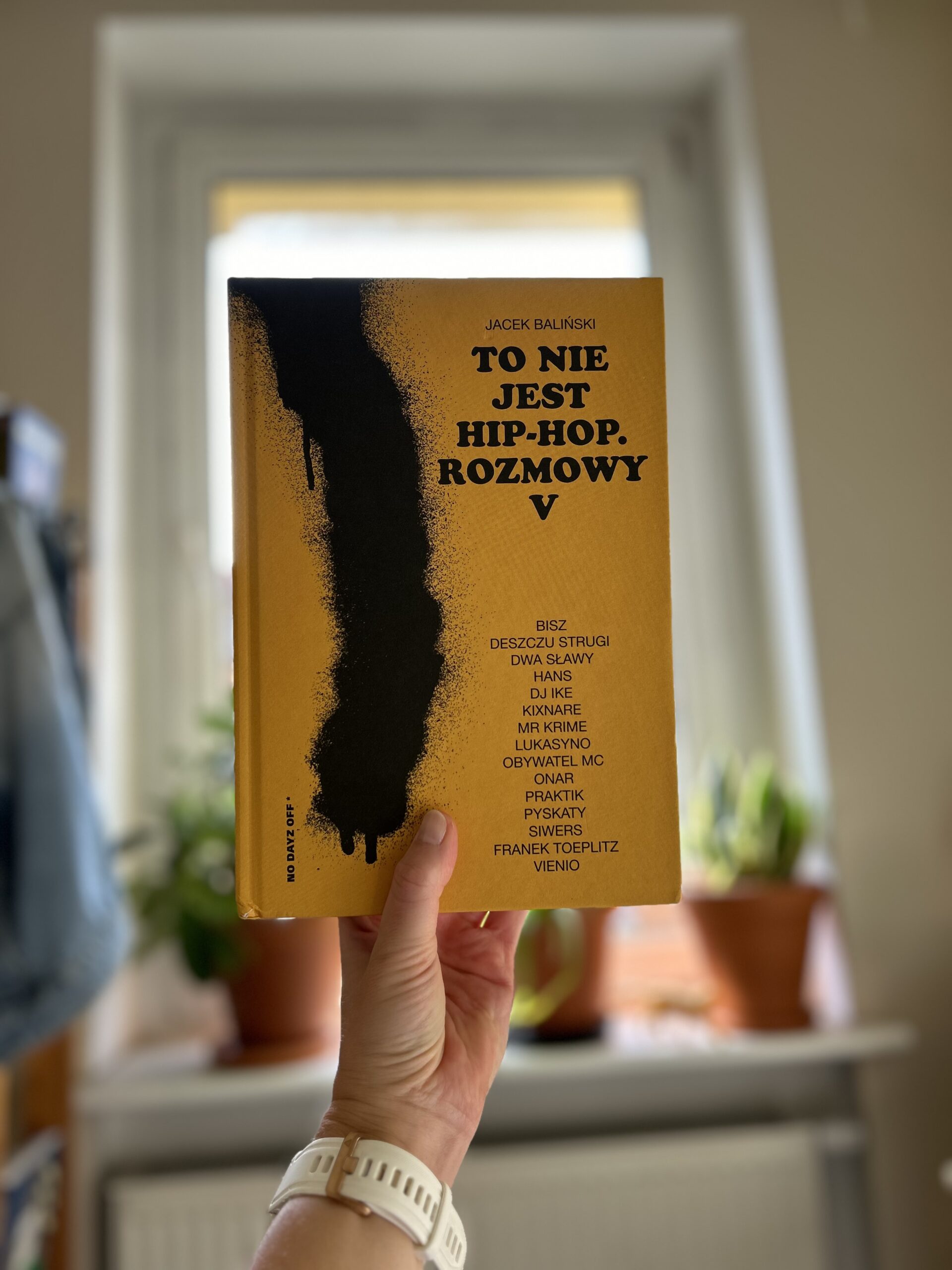 Kolorowe zdjęcie. Wyciągnięta dłoń trzyma książkę. W tle okno. Na parapecie stoją rośliny w doniczkach.