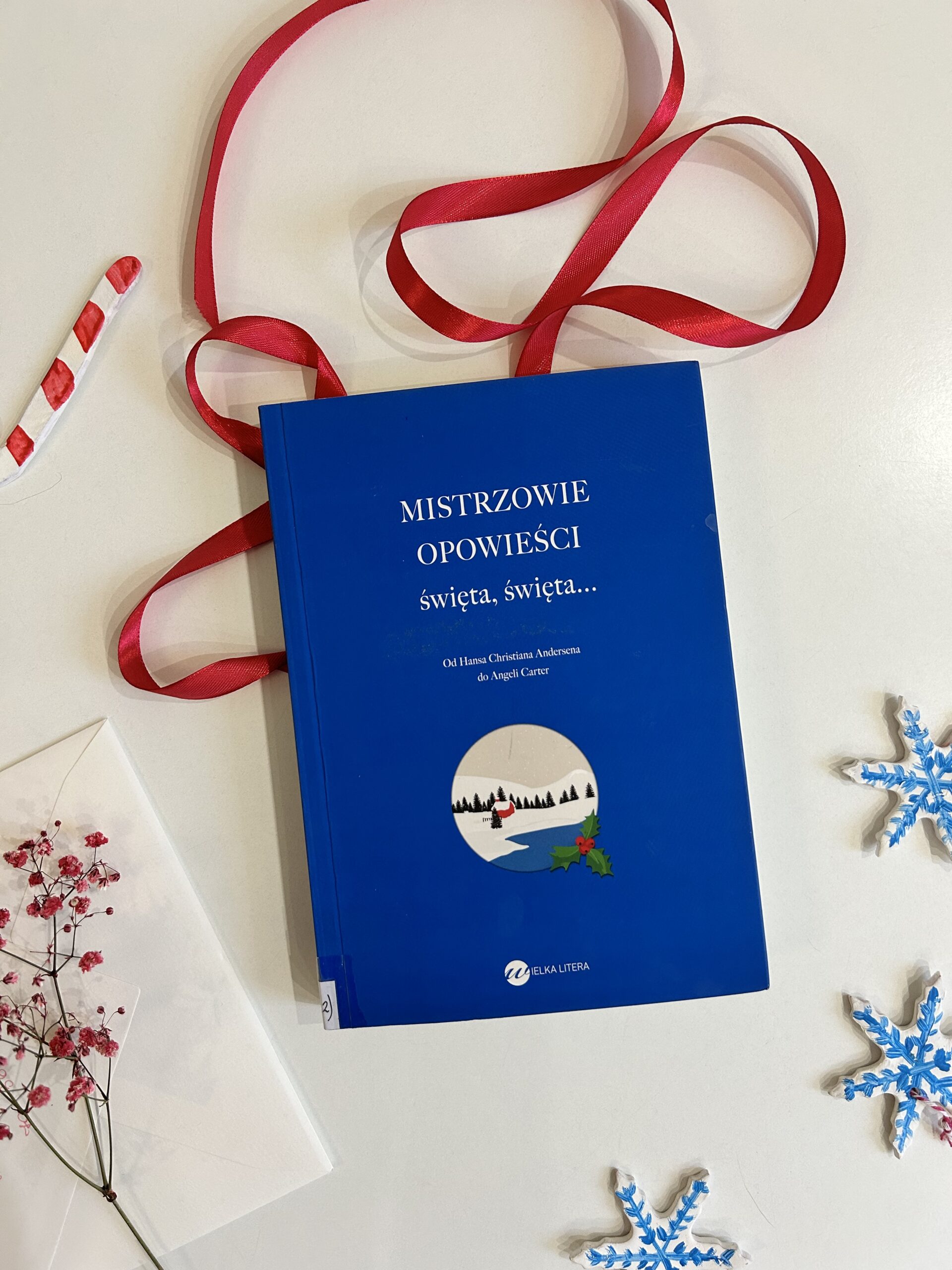 Kolorowe zdjęcie. Na białym stole leży książka. Wokół niej czerwona satynowa wstążka, gliniane zawieszki w kształcie śnieżynek i suszona czerwona gipsówka.