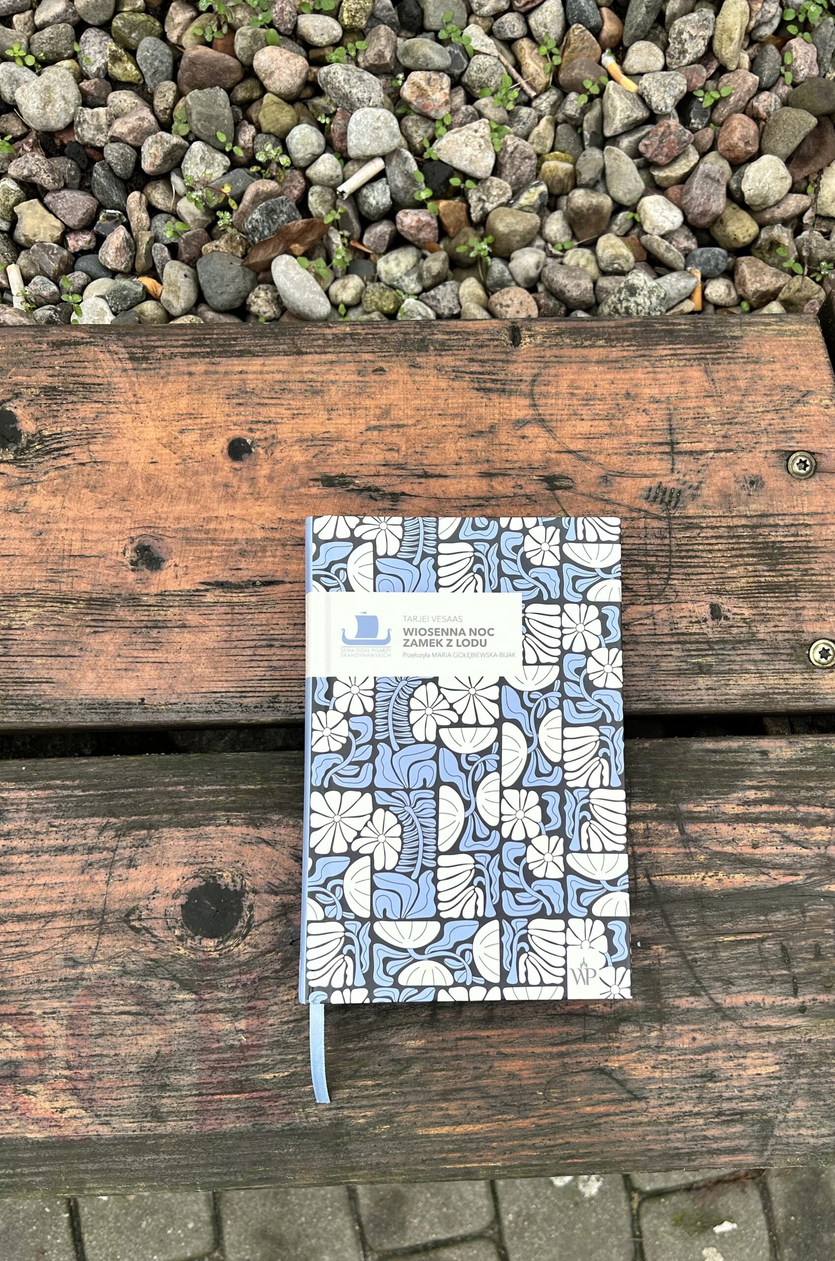 Kolorowe zdjęcie. Książka leży na drewnianej ławce. Za ławką wysypane kamienie.