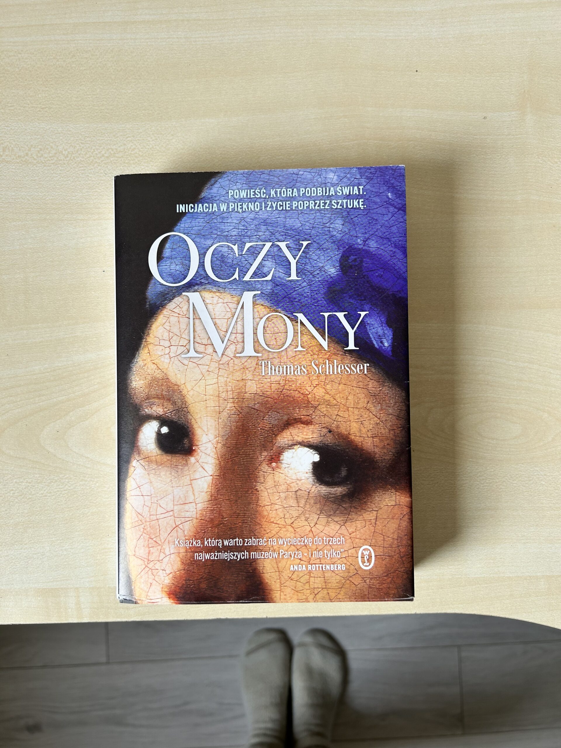 Kolorowe zdjęcie. Zamknięta książka leży na biurku. W tle stopy w wełnianych skarpetach.