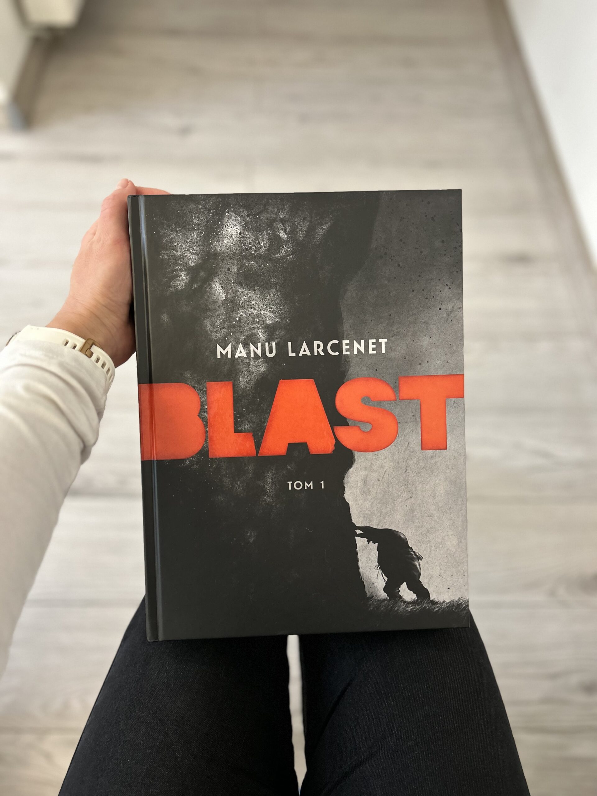 Kolorowe zdjęcie. Kobieta w wyciągniętej ręce trzyma grubą książkę. W tle drewniana podłoga.