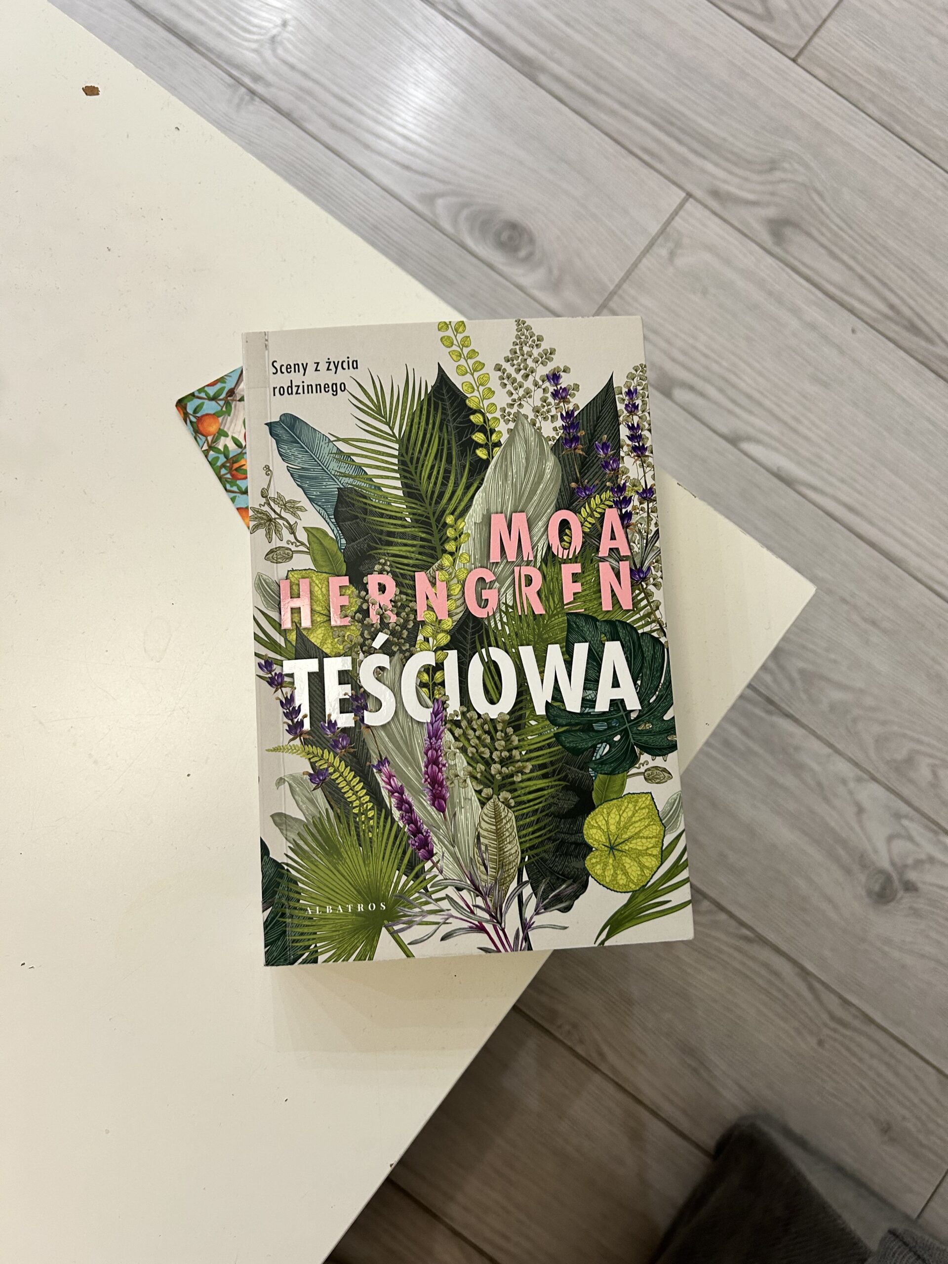 Kolorowe zdjęcie. Zamknięta książka leży na rogu stołu. Na okładce narysowane są rośliny w różnych odcieniach zieleni.