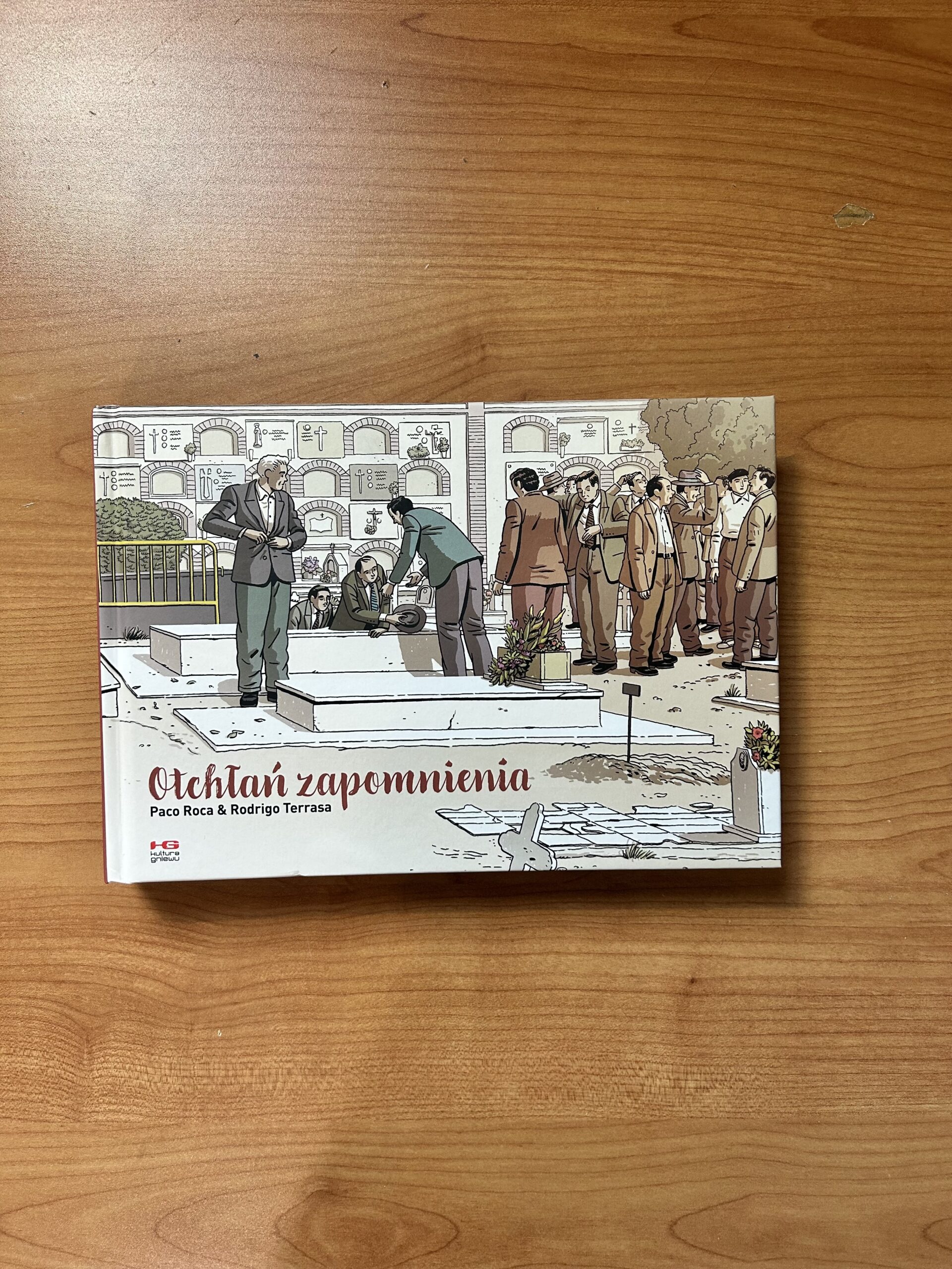 Kolorowe zdjęcie. Książka leży na drewnianym blacie. Książka jest zamknięta, ma twardą okładkę z kolorową ilustracją.