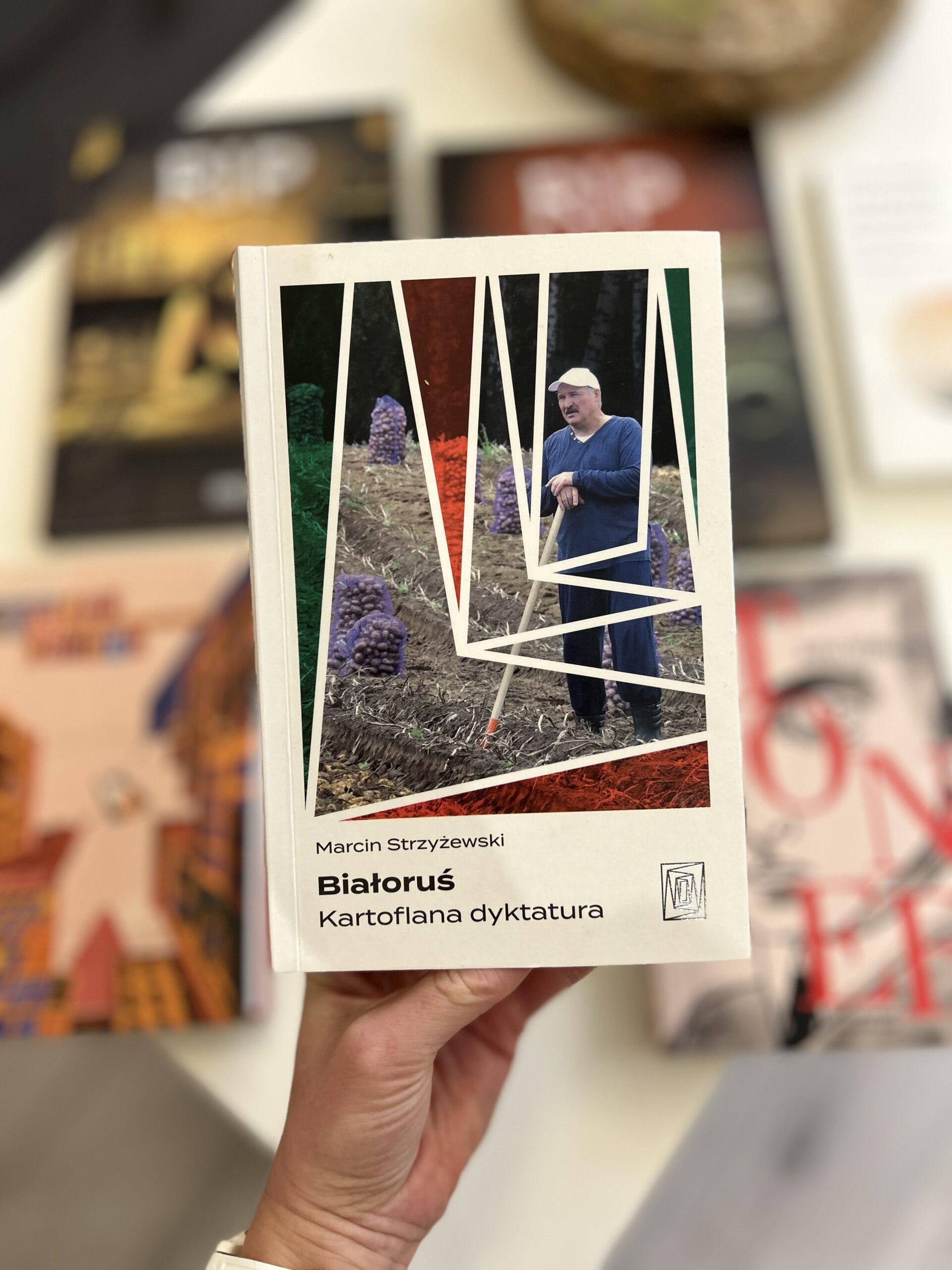 Kolorowe zdjęcie. Wyciągnięta ręka trzyma książkę. Okładka jest biała. Na środku zdjęcie Aleksandra Łukaszenki. Stoi na polu. Na nogach ma kalosze. Obok duże worki z ziemniakami.