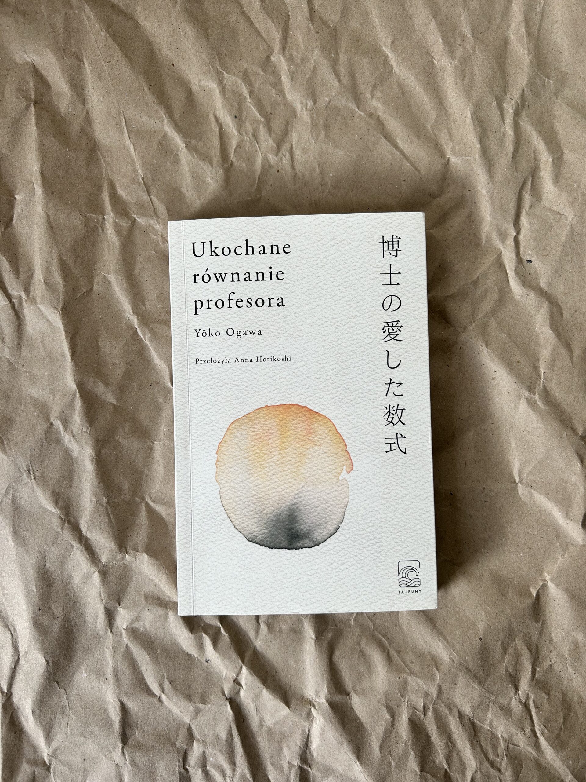 Kolorowe zdjęcie. Zamknięta książka leży na brązowym, pogniecionym papierze. Okładkę ma białą z pomarańczowo-szarą grafiką.