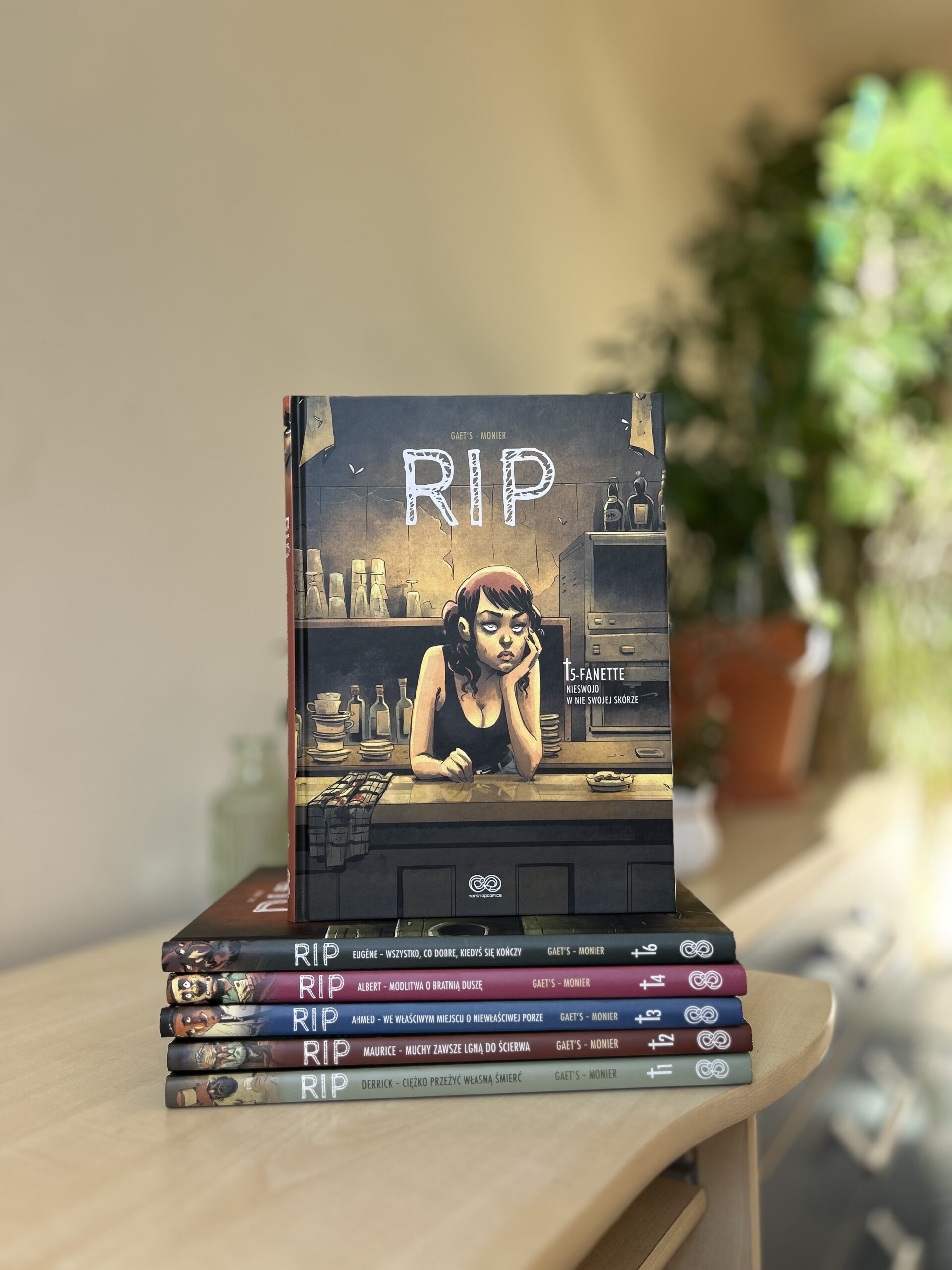 Kolorowe zdjęcie. Stos książek leży na biurku. Na szczycie jedna postawiona pionowo. W tle zielone rośliny.