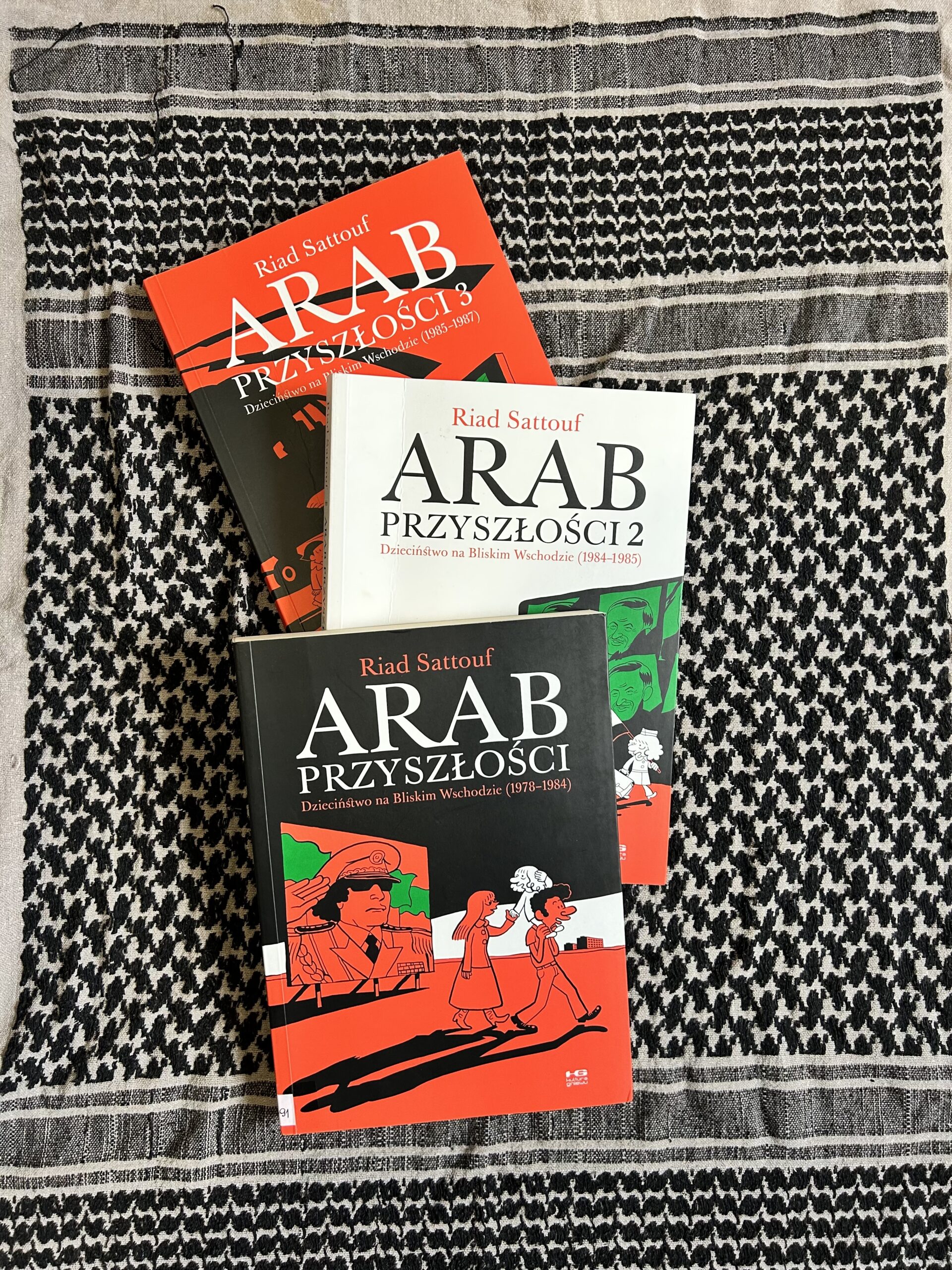 Kolorowe zdjęcie. 3 książki leżą na podłodze. Pod nimi rozłożona jest chusta. Książki mają okładki w kolorach czarny, czerwony, biały i zielony. To kolory flagi Syrii.