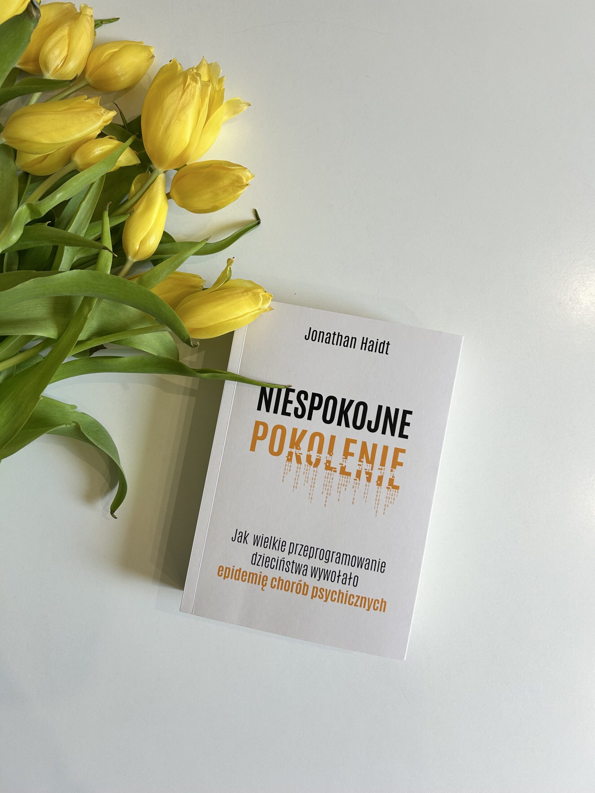 Kolorowe zdjęcie. Zamknięta książka leży na białym stole. Obok, z lewej strony, bukiet żółtych tulipanów. Okładka książki jest biała. Tytuł jest w kolorze czarnym i żółtym.