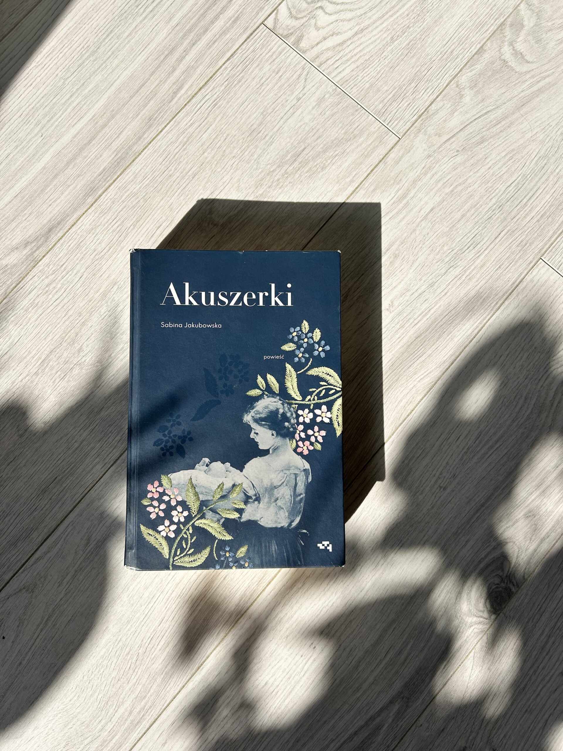 Kolorowe zdjęcie. Zamknięta książka leży na drewnianej podłodze. Padają na nią promienie słońca.