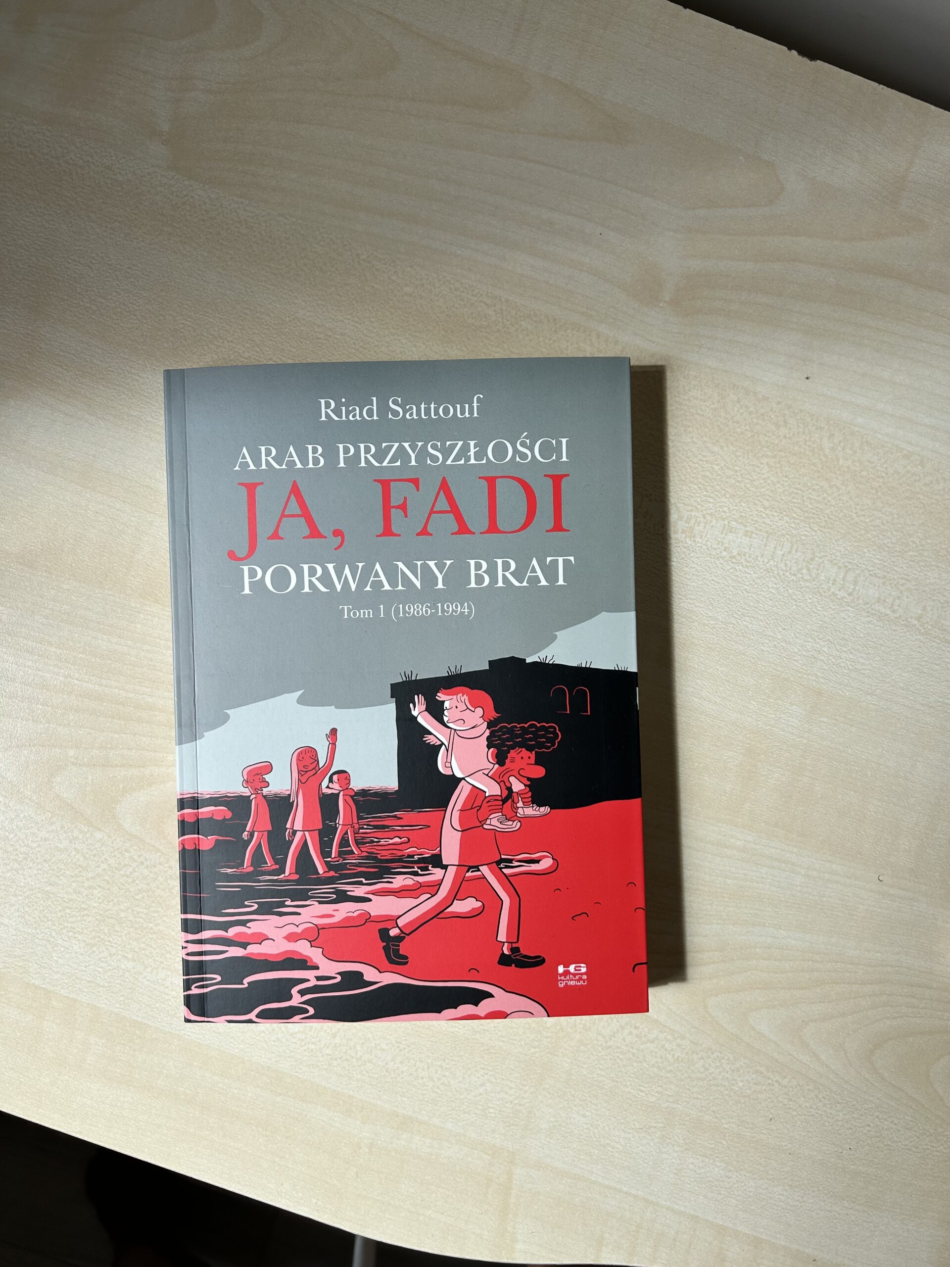 Kolorowe zdjęcie. Zamknięta książka leży na stole. Okładka jest w kolorach szarym, czarnym i czerwonym.