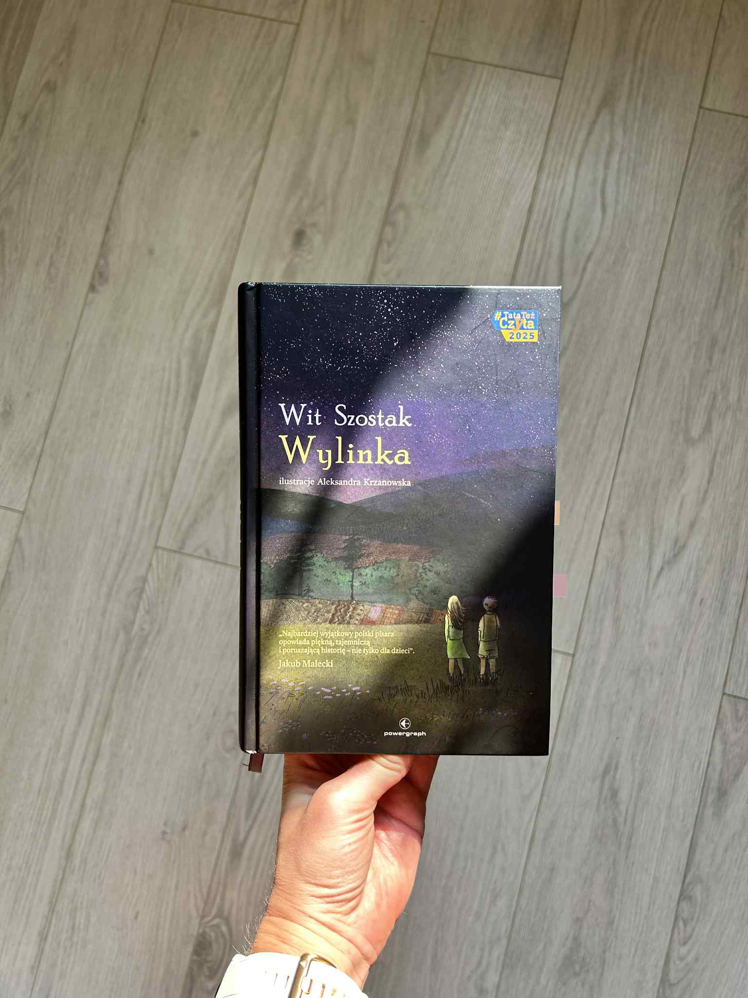 Kolorowe zdjęcie. Wyciągnięta dłoń trzyma książkę. Na książkę pada delikatne światło. Oprawiona jest w twardą, kolorową okładkę.