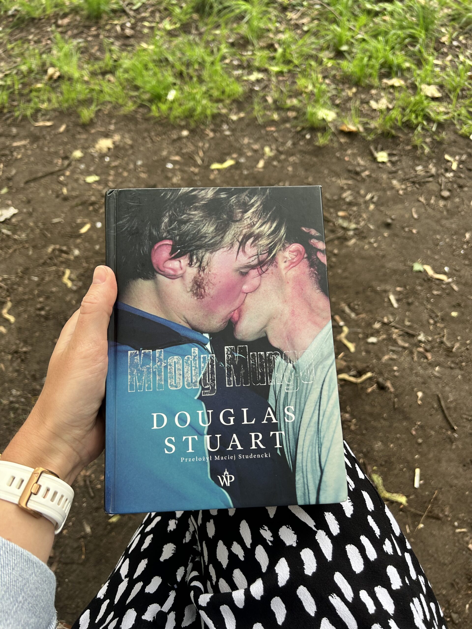 Kolorowe zdjęcie. Kobieta trzyma w lewej ręce zamkniętą książkę. W tle trawa i ziemia.