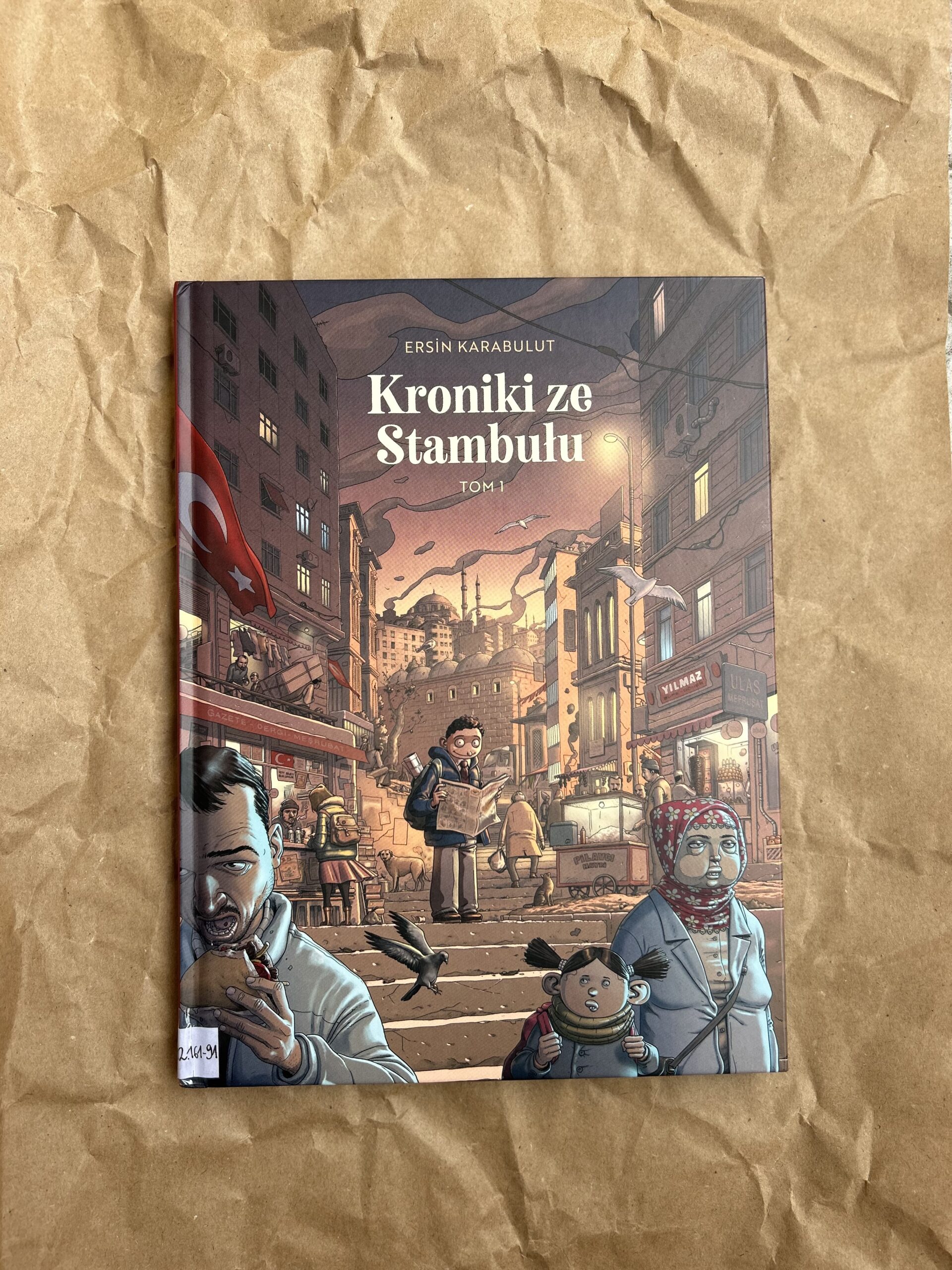 Kolorowe zdjęcie. Zamknięta książka leży na podłodze. Pod książką pognieciony, szary papier.